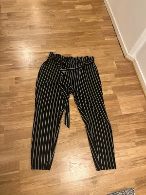 Randiga byxor från SHEIN Curve - Snygga svartvita byxor från SHEIN Curve med vita vertikala ränder och papperspåse-midja. Byxorna har avslappnad passform och resår i midjan för extra komfort. Perfekta för en trendig och bekväm stil. Strechigt tyg