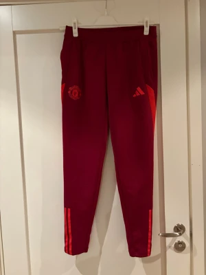 Manchester United träningsbyxor Adidas - Snygga vinröda träningsbyxor från Adidas med Manchester United-logga på ena benet och röda detaljer längs sidorna och vid bensluten. Byxorna har smal passform, dragkedja vid anklarna och elastisk midja. Perfekta för fotbollsträning eller gymmet.