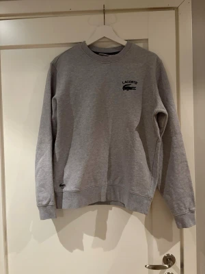 Grå sweatshirt från Lacoste - Grå sweatshirt från Lacoste med rund halsringning och broderad krokodil-logga på bröstet. Tröjan har ribbade muddar vid ärmslut och nederkant samt en avslappnad passform. Perfekt för en stilren och sportig look.