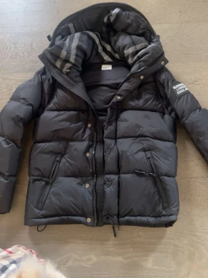 Burberry jacka o väst - Snygg svart dunjacka från Burberry med avtagbar huva och klassiskt rutigt innerfoder. Jackan har dragkedja, knappar och två snedställda fickor med dragkedja. Perfekt för kalla vinterdagar och riktigt stilren look.