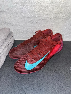 Röda Nike Mercurial fotbollsskor - Säljer ett par röda Nike Mercurial fotbollsskor med turkosa detaljer och snörning. Skorna har en smal, aerodynamisk form och är tillverkade i ett lätt syntetmaterial för bästa känsla och grepp på planen. Perfekta för dig som vill ha snabbhet och stil på fotbollsplanen.