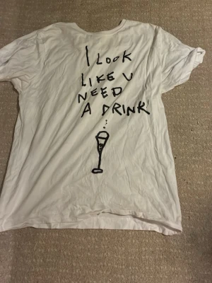 Vit t-shirt med tryck och text - Säljer en vit t-shirt med svart handskriven text på ryggen: 'I look like u need a drink' och en illustration av ett glas. T-shirten har rund hals och korta ärmar. Perfekt för dig som gillar statement-plagg med humor.
