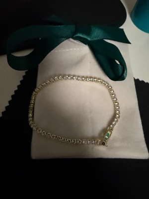 Guldigt tennisarmband med stenar - Ett elegant tennisarmband i guldton med gnistrande klara stenar runt hela bandet. Armbandet har en klassisk, smal design och stängs med ett diskret spänne. Perfekt för dig som gillar stilrena och glittriga accessoarer. Armbandet är i helt nytt skick.  OBS: kommer med mocka presentpåse på bilden! 🎁   18k gult guld pläterad, stenar är zirconia.  Top kvalitet hållbar och super snygg och glittrig.  Går att duscha med! 2,5mm tjock, 17cm lång