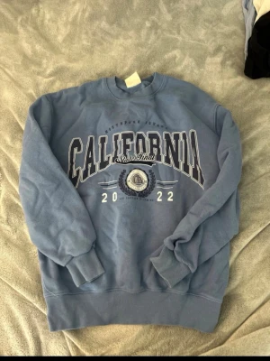 Blå sweatshirt CALIFORNIA Pull&Bear - Säljer en blå sweatshirt från Pull&Bear med stort tryck 'CALIFORNIA 2022' på bröstet och coolt emblem. Tröjan har ribbade muddar vid ärmslut och nederkant samt rund halsringning. Perfekt för en avslappnad och trendig look.