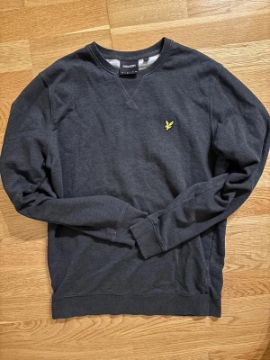 Mörkgrå sweatshirt från Lyle & Scott - Mörkgrå sweatshirt från Lyle & Scott med rund halsringning och klassisk gul logga broderad på bröstet. Tröjan har ribbade muddar vid ärmslut och nederkant, perfekt för en avslappnad och stilren look.