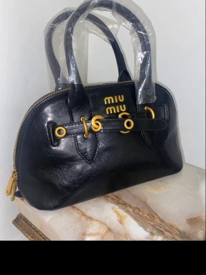 Svart handväska från Miu Miu - Snygg svart handväska från Miu Miu i glansigt läder med guldfärgade metalldetaljer och dubbla handtag. Väskan har ett stilrent, rundat formspråk och dragkedja upptill. Loggan syns tydligt framtill i guld. Perfekt för dig som gillar lyxiga accessoarer.