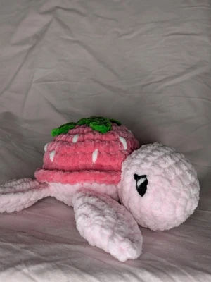Virkad sköldpaddsgosedjur jordgubb - Supergulligt handvirkat gosedjur i form av en sköldpadda med rosa kropp och ett skal som är en jordgubbe. Skalet är rosa med vita prickar och gröna blad på toppen. Sköldpaddan är 16×16 cm. Tillverkad i mjukt, fluffigt garn som känns mysigt att krama.