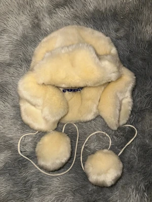 Vit pälsmössa från Polo Ralph Lauren - Säljer en fluffig vit pälsmössa från Polo Ralph Lauren med öronlappar och långa snören med stora bollar i ändarna. Mössan är fodrad med mjuk stickad ullblandning och har en lyxig vinterlook. Perfekt för dig som vill sticka ut i kylan!