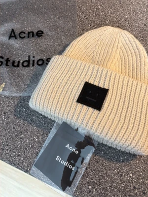  Acne Studios - Säljer en vit ribbstickad mössa från Acne Studios med uppvikt kant och det klassiska svarta ansiktslogotypen framtill. Mössan är tillverkad i mjukt material och har en clean, modern look som passar perfekt till streetwear och casual outfits.