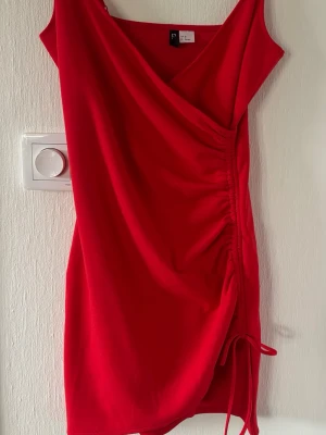 Röd omlottklänning med snörning - Snygg röd kortklänning med omlott-design och snörning i sidan. Klänningen har v-ringning och är ärmlös, vilket ger en trendig och ungdomlig look. Perfekt för dig som vill sticka ut med en färgstark outfit.