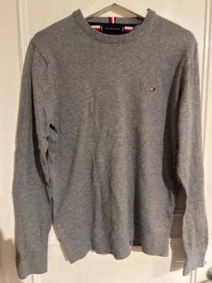 Grå stickad tröja Tommy Hilfiger - Klassisk grå stickad tröja från Tommy Hilfiger med rund halsringning och diskret logga broderad på bröstet. Tröjan har långa ärmar och ribbade muddar vid ärmslut och nederkant. Materialet är mjukt och bekvämt, perfekt för lager på lager.