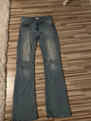 Ljusblå bootcut jeans med slitningar - Säljer ett par ljusblå bootcut jeans med slitningar över båda knäna. Jeansen har klassisk femficksdesign, knappgylf och fickor med lock bak. Tillverkade i denim med en avslappnad passform och lätt utsvängda ben. Använd fåtal gånger.