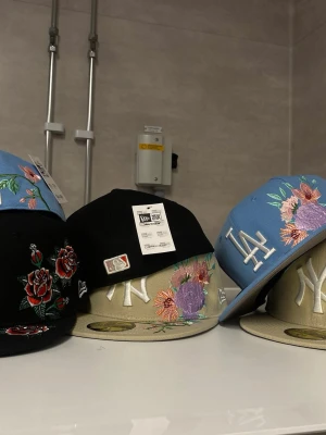 Broderade New Era kepsar med blommor(Per styck) - (DETTA ÄR PRIS PER STYCK ) Säljer flera New Era kepsar med broderade blommor och lagloggor som NY Yankees och LA Dodgers. Kepsarna finns i svart, beige och ljusblått med färgglada blommotiv och platt skärm. Perfekt för dig som vill sticka ut med en unik och trendig accessoar.