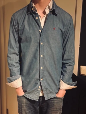 Ljusblå skjorta från Morris - Jeansaktig ljusblå skjorta från Morris med diskret röd logga broderad på bröstet. Perfekt till jeans eller chinos. Fint skick och passar M. 