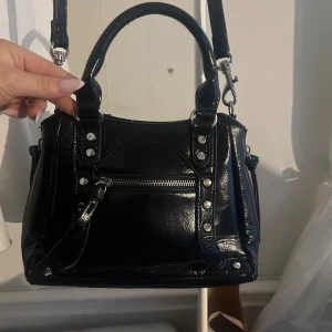 Svart handväska från NA-KD  - Snygg svart handväska från NA-KD i glansig skinnimitation med silverfärgade detaljer och dragkedja framtill. Köpt på Plick tidigare, knappt 