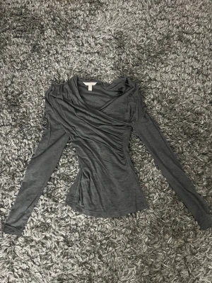 Mörkgrå omlottopp med långa ärmar - Snygg mörkgrå omlottopp med långa ärmar och figurnära passform. Toppen har draperad omlottdesign framtill och är tillverkad i ett mjukt, stretchigt material som känns skönt mot huden. Perfekt att styla med jeans eller kjol.
