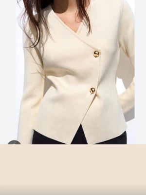 Krämvit omlottkofta med guldknappar - Säljer en stilren slutsåld krämvit omlottkofta med två stora guldfärgade knappar framtill. Koftan har lång ärm, djup v-ringning och är tillverkad i ett mjukt material som sitter snyggt på kroppen. Perfekt för dig som gillar minimalistisk och elegant stil. Storlek 38 men passar även 36