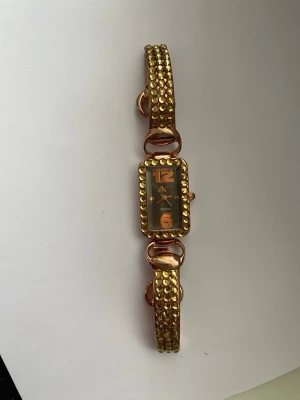 Guldig klocka med glittriga detaljer - Snygg guldig klocka med rektangulärt ur och glittriga detaljer på armbandet. Klockan har en svart urtavla med stora siffror och guldfärgade visare. Perfekt accessoar för att lyfta din outfit.