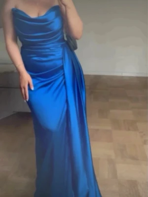 Blå axelbandslös långklänning - Säljer en elegant blå långklänning med axelbandslös design och draperad överdel. Klänningen har en figurnära passform och är tillverkad i ett glansigt material som ger en lyxig känsla. Perfekt för dig som vill sticka ut på nästa stora event!