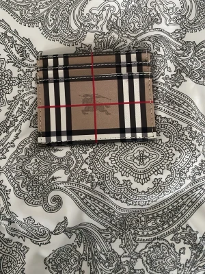 Burberry rutigt kortfodral - Snyggt kortfodral från Burberry i klassiskt beige, svart, vitt och rött rutmönster. Tillverkat i slätt läder med flera kortfack och Burberrys logga tryckt på framsidan. Perfekt för dig som vill ha en stilren och ikonisk accessoar i fickan.