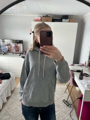 Grå hoodie med huva och ficka - Säljer en enkel och snygg grå hoodie med huva och vita snören. Tröjan har en stor magficka och ribbade muddar vid ärmslut och nederkant. Perfekt för chill dagar och passar till jeans eller joggers.