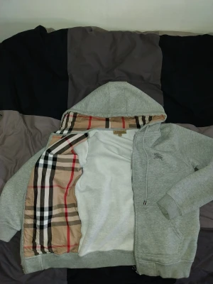 Grå zip hoodie från Burberry - Snygg grå hoodie från Burberry med dragkedja och klassiskt rutigt mönster i huvan. Hoodien har dragsko i huvan, fickor framtill och en liten Burberry-logga broderad på bröstet. Storlek S använd några gånger men inga spår av användning.