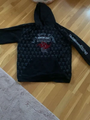 Svart hoodie med tryck och huva - Cool svart hoodie med huva och stort tryck på bröstet där det står 'Let's Party Tonight' och 'Dancefloor'. Mönster med små T:n över hela framsidan och röd detalj. Stor ficka framtill och text på ärmen, perfekt för dig som gillar streetwear