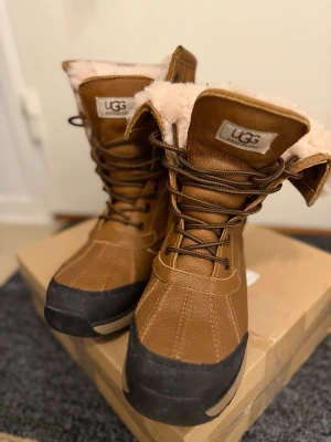 Bruna vinterstövlar från UGG - Säljer ett par bruna vinterstövlar från UGG med snörning framtill och mjukt, ljust foder. Stövlarna har en slitstark gummisula och detaljer i läder och mocka. Perfekta för kalla dagar och riktigt snygga till jeans eller leggings. Sparsamt använda. Något små i stl 