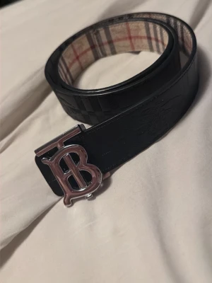 Berberry svart och beige bälte - Snyggt vändbart bälte från Berberry med klassiskt rutigt mönster på ena sidan i beige, rött och svart samt svart läder med diskret logga på andra sidan. Stort metallspänne med Burberrys ikoniska TB-logo. Perfekt accessoar för att lyfta din outfit.