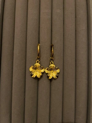 Guldpläterade örhängen med blomma - Snygga örhängen i guldpläterad metall med hängande blommor i form av fyrklöver. De har en rund creol och en detaljrik blomdesign som ger en trendig och lekfull look. Perfekta för att lyfta vilken outfit som helst.