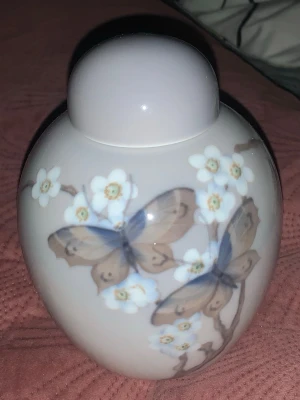 Vit porslinsburk med fjärilar och blommor - Säljer en rund vit porslinsburk med lock. Burken har ett vackert motiv av blå och bruna fjärilar samt vita blommor på grenar. Perfekt som dekoration eller förvaring av småsaker. Mjuk glansig yta och klassisk form.