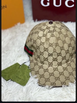 Beige keps från Gucci med GG-mönster - Snygg beige keps från Gucci med klassiskt GG-monogram över hela kepsen. Kepsen har en böjd skärm och en grön-röd rand på sidan som ger den en lyxig touch. Tillverkad i canvasmaterial och perfekt för dig som vill sticka ut.