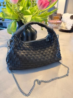 Svart flätad handväska med kedja NUNOO - Snygg svart handväska från NUNOO i flätad design med silverfärgad kedjerem och dragkedja upptill. Väskan har en mjuk, rundad form och ett kort handtag i samma material. Perfekt accessoar för att lyfta din outfit med en trendig touch.