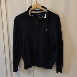 Mörkblå kofta från Polo Ralph Lauren - Snygg mörkblå tröja från Polo Ralph Lauren med dragkedja och klassisk vit broderad logga på bröstet. Kragen har vita och blå ränder som ger en snygg touch! Tröjan är långärmad och i mjuk stickad bomull passar 160-170 cm, 8/10 skick, perfekt för en stilren look✨