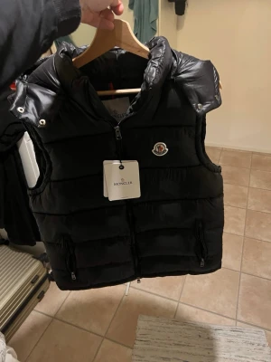 Svart dunväst från Moncler - Snygg svart dunväst från Moncler med hög krage och dragkedja framtill. Västen har två fickor med dragkedja och Moncler-logga på bröstet. Insidan har ett unikt serietidningsmärke. Perfekt för lager-på-lager och streetwear.