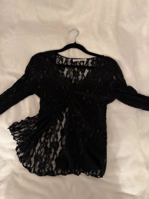 Svart spetsblus med v-ringning - Säljer en svart långärmad topp i spets med v-ringning och twistad detalj framtill. Öppen vid magen