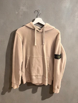 Beige hoodie från Stone Island - Säljer en beige hoodie från Stone Island med klassisk huva och dragsko. Hoodien har en stor magficka och den ikoniska svarta logotypen på vänster ärm. Perfekt för dig som gillar streetwear och enkel stil.
