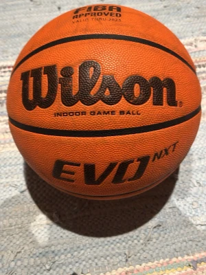 Wilson EVO NXT basketboll storlek 6 - Säljer en orange Wilson EVO NXT basketboll i storlek 6, godkänd av FIBA och märkt som indoor game ball. Bollen har svart text och detaljer samt slitstarkt greppvänligt material, perfekt för inomhusspel. Det står LG på bollen men går och ta bort med lite handsprit.