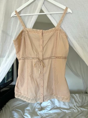 Ljusrosa spetsdetaljerad linnetopp - Söt beige linnetopp med spetsdetaljer längs kanterna och knappar framtill. Toppen har smala axelband, dekorativ knytning i midjan och fina spetsinslag både upptill och nedtill. 