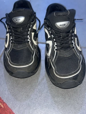 Svarta Dior sneakers med vita detaljer - Säljer ett par svarta Dior sneakers med vita och grå detaljer. Skorna har en chunky sula, meshpaneler och klassiska snörningar. Märket syns tydligt på plösen och sidan. Perfekta för dig som gillar streetwear och exklusiv design.