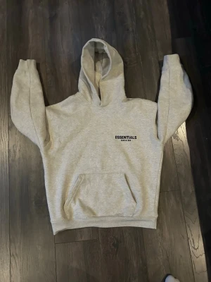 Grå Essentials hoodie  - Snygg grå hoodie från Essentials med svart tryck på bröstet. Klassisk känguruficka framtill och stor huva. Perfekt för en avslappnad streetwear-look. Mjuk bomullsblandning som är skön att ha på sig.