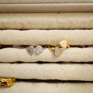 Guldfärgad ring med hjärtan - Trendig guldfärgad ring med öppen design och två hjärtformade detaljer. Ena hjärtat är slätt medan det andra är dekorerat med små gnistrande stenar för extra bling. Perfekt accessoar för dig som gillar romantiska och stilrena smycken.