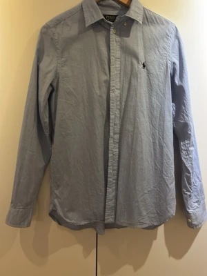 Ljusblå skjorta från Polo Ralph Lauren - Klassisk ljusblå skjorta från Polo Ralph Lauren med knappstängning och broderad logga på bröstet. Skjortan har lång ärm, normal passform och är tillverkad i bomull. Perfekt för en clean och stilren look.