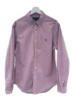 Rutig skjorta från Ralph Lauren - Klassisk rutig skjorta från Ralph Lauren i rosa, blått och vitt. Skjortan har lång ärm, knappar framtill och button-down krage. Broderad logga i blått på bröstet. Tillverkad i mjuk bomull för en bekväm känsla.