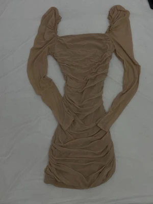 Beige långärmad kortklänning - Trendig beige kortklänning med långa ärmar och rynkade detaljer längs hela klänningen. Klänningen har en figurnära passform och är tillverkad i ett mjukt, stretchigt material som ger en snygg siluett.