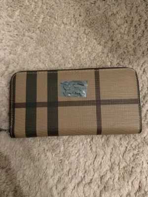 Burberry plånbok i klassiskt rutmönster - Snygg plånbok från Burberry i beige och brunt med det ikoniska rutmönstret och silverfärgad logga framtill. Plånboken har dragkedja runt om och är tillverkad i tåligt canvasmaterial. Perfekt för dig som gillar stilrena accessoarer med lyxig känsla.