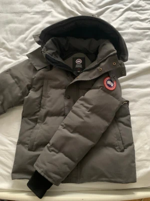 Grå pufferjacka från Canada Goose - Snygg grå pufferjacka från Canada Goose med ikonisk logotyp på ärmen. Jackan har stor huva, dragkedja och knappar framtill samt två sidofickor. Tjockt vadderad för maximal värme och har ribbade muddar vid ärmsluten. Perfekt för kalla vinterdagar. 