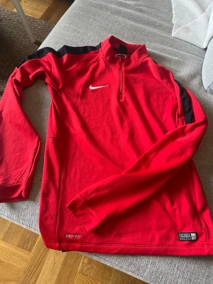 Röd Nike Dri-Fit träningströja - Säljer en röd långärmad träningströja från Nike med Dri-Fit material som håller dig torr under träningen. Tröjan har en halv dragkedja framtill, svarta detaljer på axlarna och Nike-logga på bröstet. Perfekt för fotboll eller annan sport. Storlek S men mer som M.