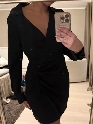 Svart omlottklänning med v-ringning - Stilren svart omlottklänning med v-ringning och långa ärmar. Klänningen har en figurnära passform och lätt drapering i midjan som ger en snygg siluett. Perfekt för dig som gillar en klassisk och elegant look.