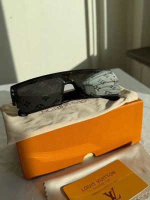 Svarta Louis Vuitton monogram solglasögon - Säljer ett par svarta solglasögon från Louis Vuitton med ikoniskt monogrammönster på glaset. Glasögonen har en futuristisk, rektangulär form och breda skalmar. Snygg accessoar för dig som vill sticka ut och addera lyx till din stil.